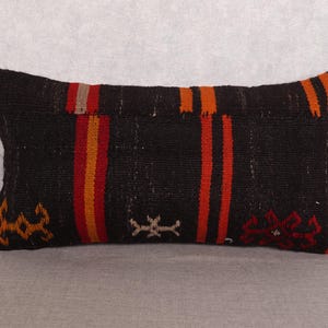Op de afbeelding: Rechthoekig zwart kussen met verticale oranje en rode strepen. Het kussen heeft een geweven textuur en heeft kleine decoratieve patronen in oranje, rood en gebroken wit. Het ligt op een grijze ondergrond.