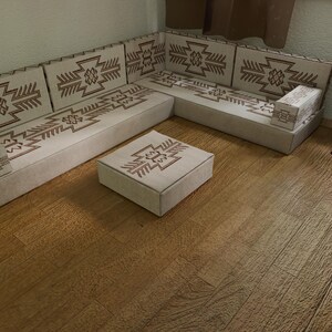 May include: A beige sectional sofa and matching ottoman with brown geometric patterns. The sofa is arranged in an L-shape, with a small round side table and a potted plant nearby. The floor is made of wood.