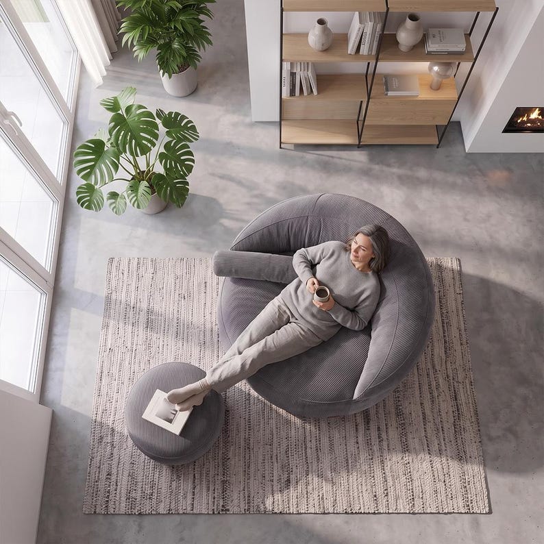 May include: A large, round, grey corduroy lounge chair with a matching cylindrical bolster pillow and a circular ottoman. The chair features a textured fabric surface and a deep, curved seat design, providing a comfortable space for relaxation.