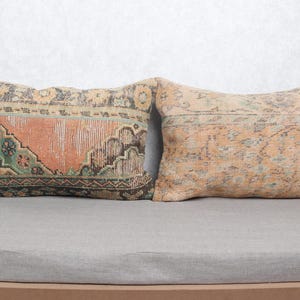 Op de afbeelding: Twee rechthoekige kussens met een vintage tapijtontwerp. Eén kussen heeft een oranje middenpaneel met groene en zwarte randen. Het andere kussen heeft een vervaagd oranje en beige patroon. Beide kussens liggen op een grijs oppervlak.