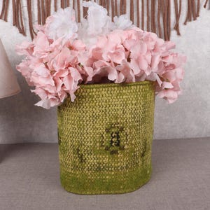 May include: A green woven basket, filled with pink and white flowers. The basket has a textured finish and is cylindrical. A wooden lamp with a beige lampshade is to the left of the basket.