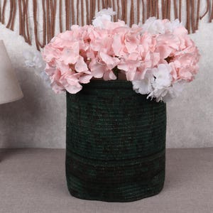 Puede incluir: Una cesta tejida de color verde oscuro llena de flores artificiales rosas y blancas. La cesta tiene forma cilíndrica y está colocada sobre una superficie de color neutro. Una lámpara de madera con una pantalla beige es visible en el fondo.