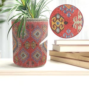 Könnte beinhalten: Ein zylindrischer Blumentopf mit einem roten Stoffmuster. Das Muster umfasst geometrische Formen in Blau, Gelb und Braun. Der Topf enthält eine grüne Pflanze mit langen, dünnen Blättern. Eine Nahaufnahme zeigt die Textur des Stoffes.