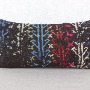 May include: Rectangular decorative pillow with a dark brown background and a colorful, patterned design. The pattern includes blue, red, white, and pink tree-like shapes. The pillow is likely made of a woven fabric.
