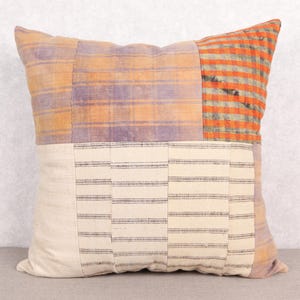 May include: Square cushion with a patchwork design. The cushion features sections of orange and purple plaid, and a cream-coloured section with horizontal black stripes. The cushion is a square shape and appears to be made of fabric.