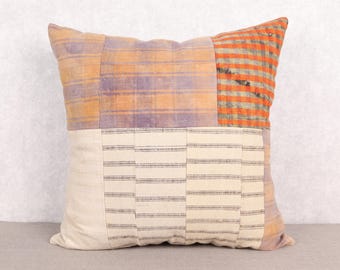 May include: Square decorative pillow with a patchwork design. The pillow features sections of orange and purple plaid, and a cream-colored section with horizontal black stripes. The pillow is a square shape and appears to be made of fabric.