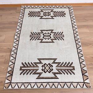 May include: Rectangular rug with a light gray background and brown geometric patterns. The rug features three central designs and a border with a zigzag pattern. The rug is placed on a wooden floor.