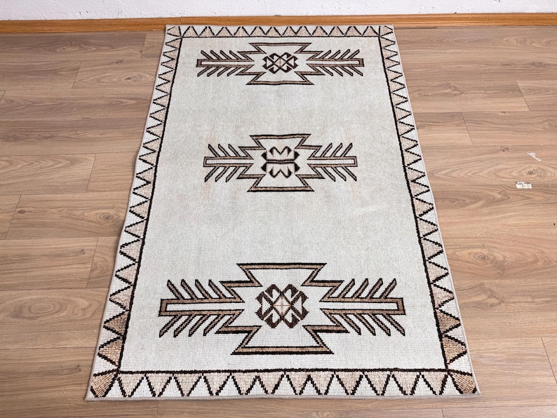 May include: Rectangular rug with a light gray background and brown geometric patterns. The rug features three central designs and a border with a zigzag pattern. The rug is placed on a wooden floor.