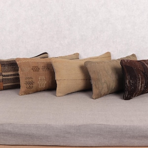 Puede incluir: Cinco cojines rectangulares en tonos marrones y beige están dispuestos sobre una superficie gris. Los cojines tienen varios patrones, incluyendo rayas y diseños geométricos, y parecen estar hechos de tela tejida.