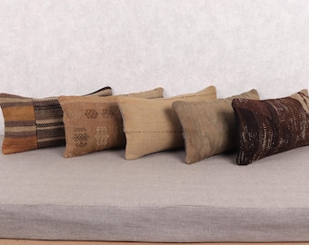 May include: Five rectangular throw pillows in shades of brown and tan are arranged on a gray surface. The pillows have various patterns, including stripes and geometric designs, and appear to be made of woven fabric.