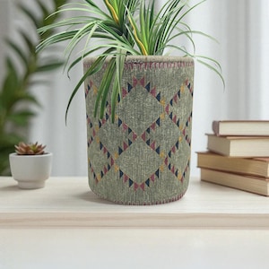 May include: A cylindrical plant pot with a green fabric exterior. The fabric is patterned with a repeating diamond design of red, yellow, and navy blue triangles. A green plant with long, thin leaves is inside the pot.
