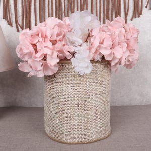May include: A woven basket filled with pink and white artificial flowers. The basket is cylindrical and has a textured, beige appearance. A wooden lamp with a beige lampshade is to the left of the basket.