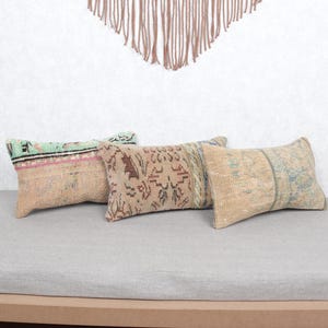 May include: Three rectangular cushions with patterned designs in beige, brown, and teal hues. The cushions are arranged on a grey surface, with a decorative wall hanging in the background. The cushions appear to be made of a woven fabric.