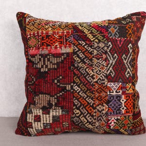 May include: Square decorative pillow with a patchwork design. The pillow features a mix of geometric patterns in shades of red, brown, orange, and purple. The pillow is likely made of woven fabric.
