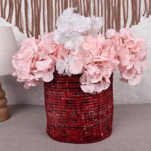 Puede incluir: Una cesta roja tejida llena de flores artificiales rosas y blancas. A la izquierda hay una lámpara de madera con una pantalla beige. El fondo incluye una pared texturizada y un tapiz de pared tejido marrón.
