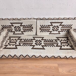 May include: A cream-colored, modular sofa with geometric patterns in brown. The sofa includes a base, two back cushions, and two armrests. The design features repeating geometric shapes and a zig-zag border along the base.