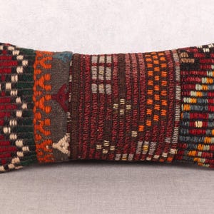 May include: Rectangular decorative pillow with a patchwork design. The pillow features sections of woven fabric in various colors, including red, green, orange, and brown. The pillow is placed on a neutral gray surface.