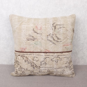 May include: Square beige cushion with a vintage, distressed appearance. It has a two-tone design, with a darker brown band separating the top and bottom. The top features faded floral patterns, and the bottom has geometric designs.