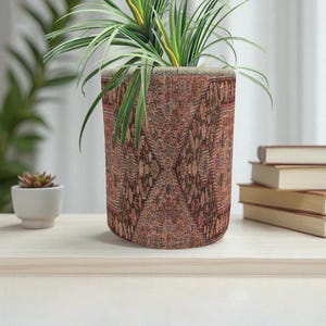 Könnte beinhalten: Ein zylindrischer Blumentopf mit einem detaillierten, gemusterten Design in Braun-, Rot- und Beigetönen. Der Topf enthält eine grüne Pflanze mit langen, schlanken Blättern. Der Topf steht auf einer hellen Oberfläche.