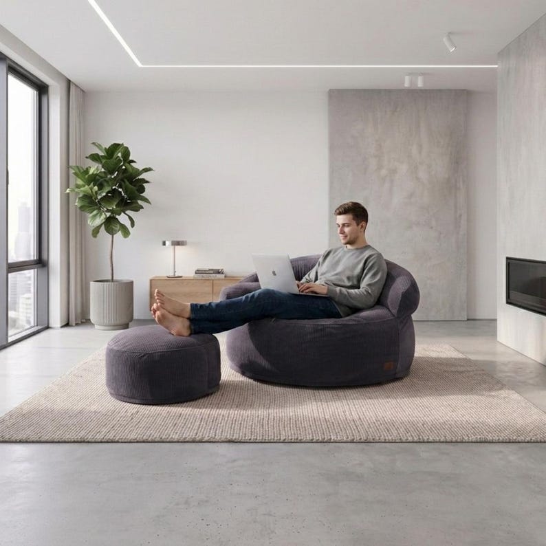 May include: A dark gray corduroy bean bag chair with a matching circular ottoman. The chair features a rounded, low-profile design with soft, textured fabric, providing a comfortable seat for working on a laptop in a modern living space.