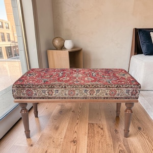 Puede incluir: Banco rectangular con una parte superior tapizada con estampado rojo y beige. El banco tiene un marco de madera y cuatro patas torneadas. El patrón de la parte superior presenta diseños florales y geométricos. El banco está frente a una ventana.