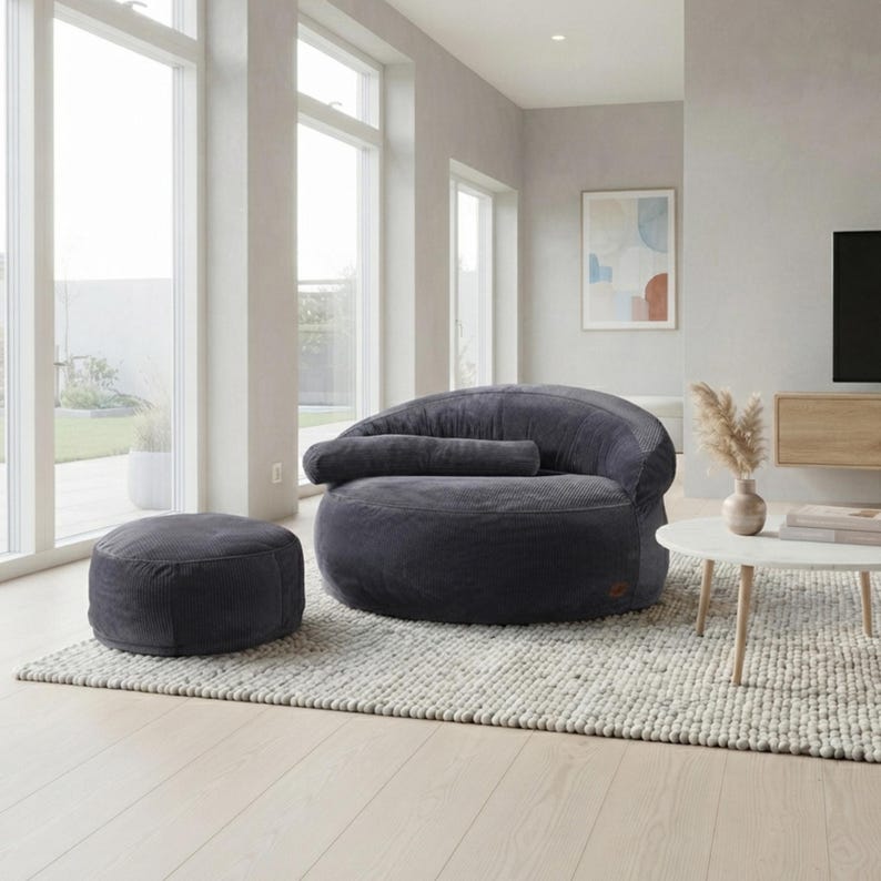 May include: A dark gray corduroy bean bag chair with a matching round ottoman. The chair features a low-profile, rounded silhouette with a horizontal bolster cushion across the backrest, providing a soft, textured seating option for a living space.