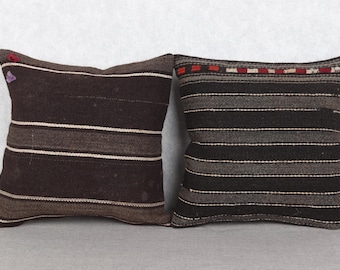 May include: Two square throw pillows with a woven texture. One pillow is primarily dark brown with horizontal white stripes. The other is dark gray with horizontal stripes and a decorative top border with red and white accents. Both pillows are on a gray surface.