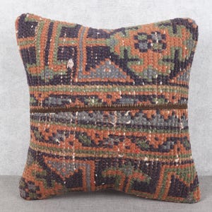 May include: Square decorative pillow with a woven, geometric pattern in shades of orange, green, gray, and dark purple. The pillow has a textured appearance and a brown zipper.