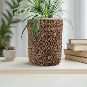 Puede incluir: Una maceta cilíndrica con un patrón detallado y ornamentado en tonos marrón y dorado. La maceta contiene una planta verde con hojas largas y delgadas. La maceta está sobre una superficie clara.