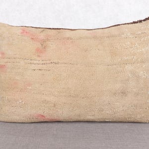 May include: Rectangular beige pillow with a textured surface. The pillow has horizontal lines and small red accents. The edges are a darker brown color. This decorative pillow is suitable for a living room or bedroom.