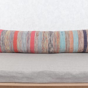 Puede incluir: Un largo cojín rectangular con un estampado de rayas en tonos rojos, naranjas, azules y grises. El cojín está colocado sobre una superficie gris, y el fondo es una pared blanca lisa. El cojín mide aproximadamente 91 cm de largo.