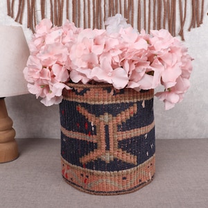 May include: A cylindrical woven basket with a geometric pattern in navy blue, beige, and red holds a bouquet of pink flowers. A wooden lamp with a white lampshade is to the left. The background is a neutral color.