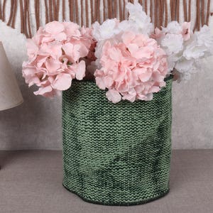 Puede incluir: Un jarrón verde tejido contiene flores rosas y blancas. El jarrón tiene una apariencia texturizada y una forma cilíndrica. A la izquierda del jarrón hay una lámpara de madera con una pantalla beige.