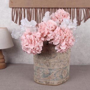 May include: A decorative arrangement of pink and white artificial hydrangeas in a woven basket. A small wooden lamp with a beige lampshade is to the left. A brown wall hanging with fringe is in the background.