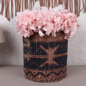 Puede incluir: Una cesta tejida cilíndrica con un patrón geométrico marrón y negro, llena de flores rosas y blancas. La cesta está sobre una superficie neutra. A la izquierda hay una lámpara de madera con una pantalla beige.