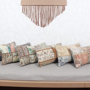 May include: Six rectangular throw pillows with patchwork designs in various colors, including beige, blue, and pink, are arranged on a gray surface. The pillows have a vintage, textured appearance. A decorative wall hanging and a vase are in the background.