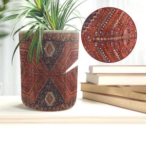 May include: A cylindrical plant pot with a detailed, patterned design in shades of red, brown, and navy blue. The pot holds a green plant with long, slender leaves. A close-up shows the intricate woven texture of the pot's fabric.
