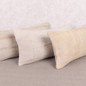 Puede incluir: Tres cojines rectangulares en tonos beige y crema. Los cojines tienen un aspecto texturizado y tejido, con costuras visibles y sutiles variaciones de color. Los cojines están colocados sobre una superficie de tono neutro.