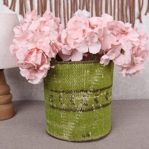 May include: A green woven vase filled with pink flowers. The vase has a dark brown band around the middle. A wooden lamp with a white lampshade is to the left of the vase.