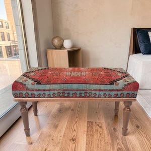 Puede incluir: Un otomán rectangular con una parte superior estampada en rojo, azul y beige. El marco de madera tiene cuatro patas torneadas marrones. El otomán se coloca frente a una ventana, con una pared de tonos neutros y suelo de madera.