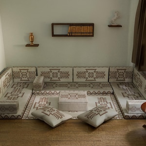 Puede incluir: Una zona de asientos con patrones beige y marrón, cojines a juego y una mesa de centro central. La habitación cuenta con una pequeña mesa auxiliar redonda, una ventana y estantes decorativos en la pared. El diseño general sugiere un tema cultural y acogedor.