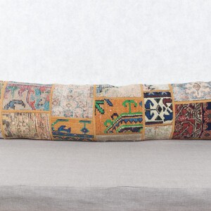 May include: A long, rectangular pillow with a patchwork design. The pillow features various rectangular patches of different colors and patterns, including red, blue, yellow, and green. The pillow is placed on a gray surface.