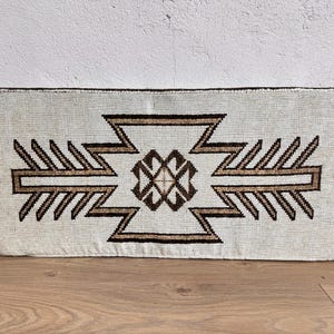 May include: Rectangular off-white cushion with a geometric pattern in shades of brown. The design features a central diamond shape with zigzag and arrow-like elements. The cushion is placed on a wooden surface.