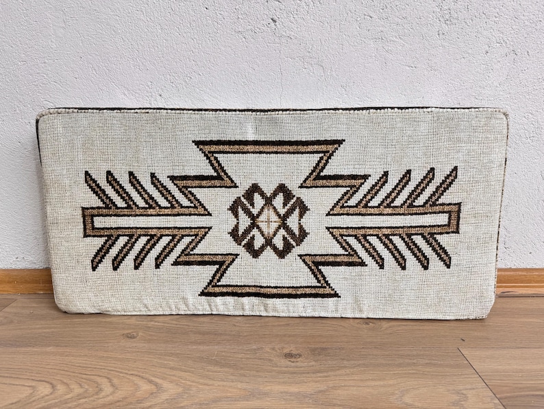 May include: Rectangular off-white cushion with a geometric pattern in shades of brown. The design features a central diamond shape with zigzag and arrow-like elements. The cushion is placed on a wooden surface.