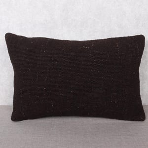 Può includere: Un cuscino rettangolare marrone scuro con una superficie strutturata. Il cuscino è realizzato in materiale tessuto e si trova su una superficie grigia. Un elemento decorativo.