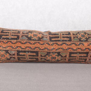 May include: Rectangular decorative pillow with a geometric pattern in shades of brown, orange, and green. The pillow has a textured appearance and is placed on a gray surface. The pillow's design features repeating shapes and lines.