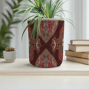 Peut inclure: Un pot de plante cylindrique avec un motif dans les tons de marron, rouge et beige. Le pot contient une plante verte avec de longues feuilles fines. Le pot est posé sur une surface en bois de couleur claire.