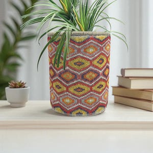 May include: A cylindrical plant pot with a colorful geometric pattern in red, yellow, and orange. The pot holds a green plant with long, slender leaves. The pot sits on a light-colored wooden surface.