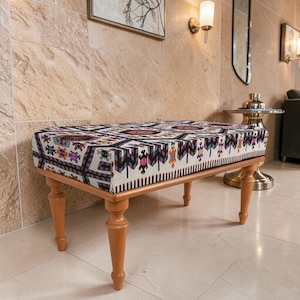 May include: A rectangular bench with a patterned fabric top and wooden legs. The fabric features a geometric design in shades of red, blue, pink, and cream. The bench has four turned legs and is set against a neutral-toned wall.