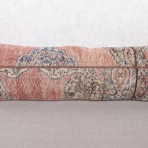 May include: A long, rectangular pillow with a vintage rug design. The pillow features a patchwork of faded red, blue, and cream-colored patterns. The pillow is approximately 36 inches long.